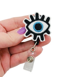 May include: A black and white eye with a blue pupil on a white background. The eye is attached to a clear plastic retractable badge holder.