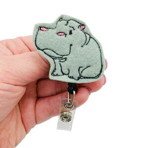 May include: A light blue felt hippopotamus badge reel with pink ears and black eyes. The reel has a black cord and a clear plastic clip.