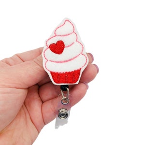 May include: White and red felt cupcake with a red heart on top, attached to a black and clear retractable badge reel.