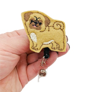 May include: A tan and brown felt pug dog badge reel. The pug has embroidered details, including a black nose and eyes, and a pink tongue. The badge reel has a black beaded cord and a clear plastic ID holder.