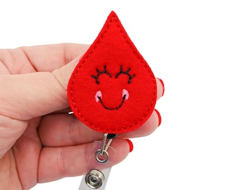 May include: Red felt blood drop with a smiling face, black eyelashes, and pink cheeks. The blood drop is attached to a clear plastic retractable badge holder with a black clip.