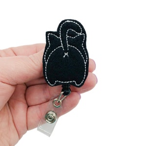 May include: A black felt badge reel featuring a cat's rear end design with white stitching. The badge reel has a retractable cord and a clear plastic snap-on holder for attaching to clothing or a lanyard.