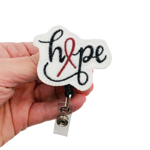 May include: White felt badge holder with a black embroidered "hope" with a red ribbon. The badge holder has a black retractable badge reel.