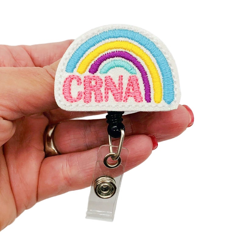 May include: A white rainbow with yellow, blue, purple, and teal stripes with the word "CRNA" in pink lettering. The rainbow is attached to a black retractable badge reel with a clear plastic clip.
