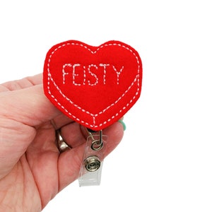Puede incluir: Un portatarjetas en forma de coraz&oacute;n de fieltro rojo con la palabra "FEISTY" bordada en blanco. El portatarjetas tiene un borde cosido blanco y un clip de pl&aacute;stico transparente. El portatarjetas se sostiene en una mano.