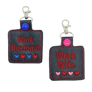 May include: Two black leather keychains with silver key rings and red embroidered text. One keychain says "Work Husband" with blue hearts and the other says "Work Wife" with pink hearts.