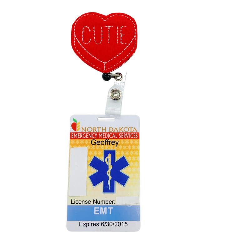 May include: Red felt heart with the word "cutie" stitched on it, attached to a white retractable badge holder with a blue and white star of life badge that says "North Dakota Emergency Medical Services" and "Geoffrey" with the text "License Number: EMT Expires 6/30/2015"