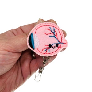 May include: A pink felt eye anatomy pin with red and blue embroidery. The pin is attached to a silver metal clip.
