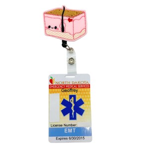 May include: A pink and brown cartoon badge reel with a cute face and a red heart. The badge reel is attached to a white plastic badge holder with the text "NORTH DAKOTA EMERGENCY MEDICAL SERVICES Geoffrey License Number: EMT Expires 6/30/2015".