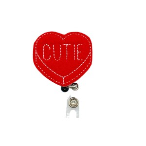 May include: Red felt heart-shaped badge holder with white stitching and the word "CUTIE" embroidered on it. The badge holder is attached to a retractable badge reel.