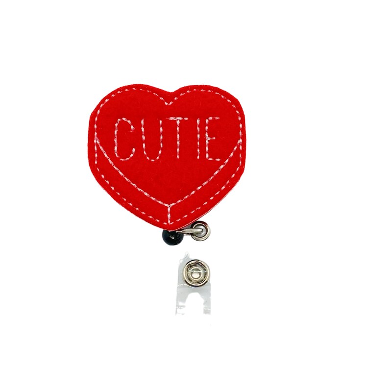 May include: Red felt heart-shaped badge holder with white stitching and the word "CUTIE" embroidered on it. The badge holder is attached to a retractable badge reel.