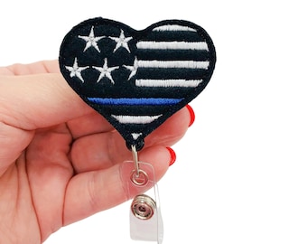 May include: A black and white heart-shaped badge holder with a thin blue line and stars. The badge holder is attached to a clear retractable badge reel.