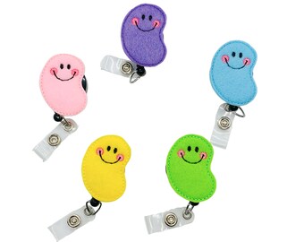 May include: Five colorful felt bean-shaped retractable badge holders with smiling faces. The holders are purple, pink, blue, yellow, and green.