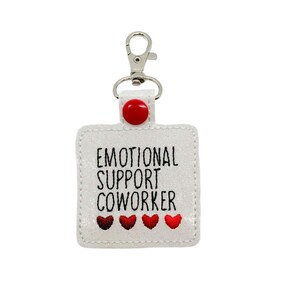 May include: White fabric keychain with a silver clip and a red button. The keychain has black embroidered text that reads "Emotional Support Coworker" with three red hearts below the text.