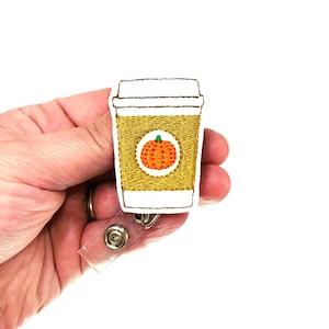 May include: A white and gold embroidered badge reel featuring a coffee cup with a pumpkin inside. The coffee cup is filled with a light brown liquid. The badge reel is attached to a clear plastic retractable cord.