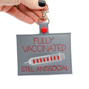 May include: Gray fabric key chain with a red button and a silver clip. The key chain has red embroidered text that reads "FULLY VACCINATED STILL ANTISOCIAL" and a white embroidered image of a syringe.