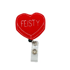Puede incluir: Un portatarjetas rojo en forma de coraz&oacute;n con la palabra "FEISTY" bordada en blanco. El portatarjetas tiene un borde cosido blanco y un clip de pl&aacute;stico transparente. El carrete est&aacute; sujeto a un cable retr&aacute;ctil negro.