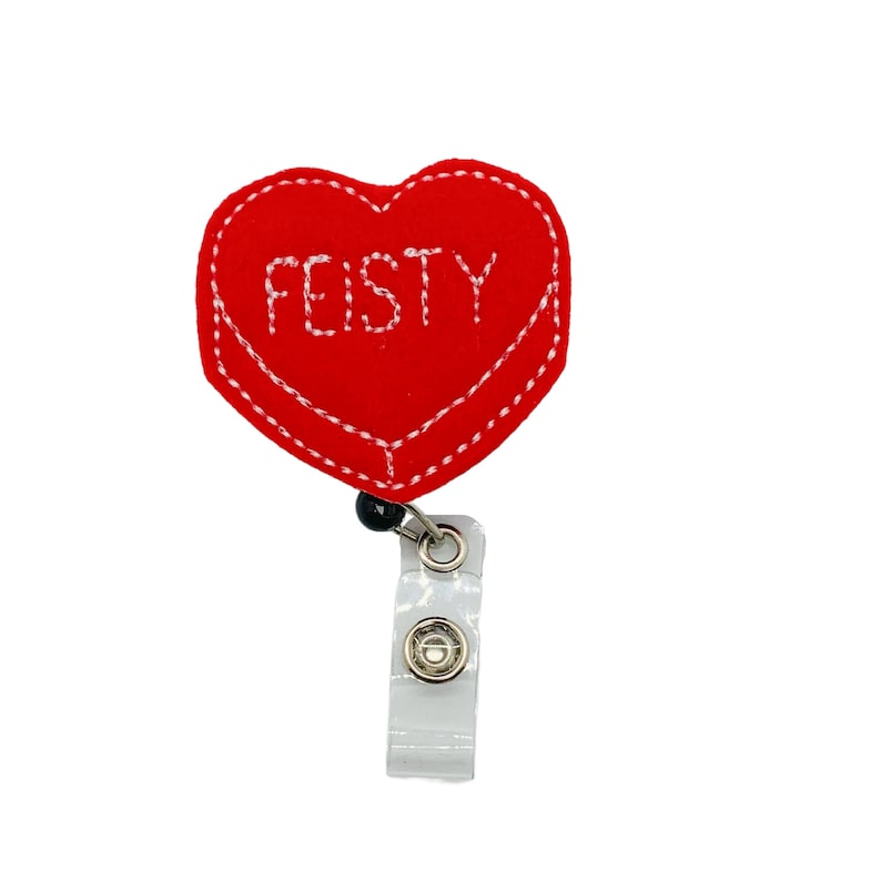 Puede incluir: Un portatarjetas rojo en forma de coraz&oacute;n con la palabra "FEISTY" bordada en blanco. El portatarjetas tiene un borde cosido blanco y un clip de pl&aacute;stico transparente. El carrete est&aacute; sujeto a un cable retr&aacute;ctil negro.
