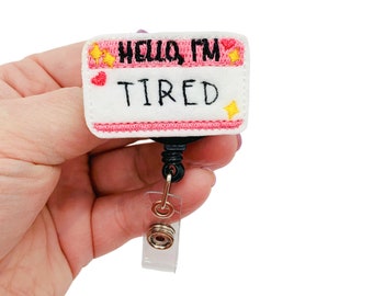 以下が含まれることがあります： 黒色の刺繍で「Hello, I'm Tired」と書いてあるピンクと白のネームタグ。ネームタグは黒いリトラクタブルバッジホルダーに取り付けられています。