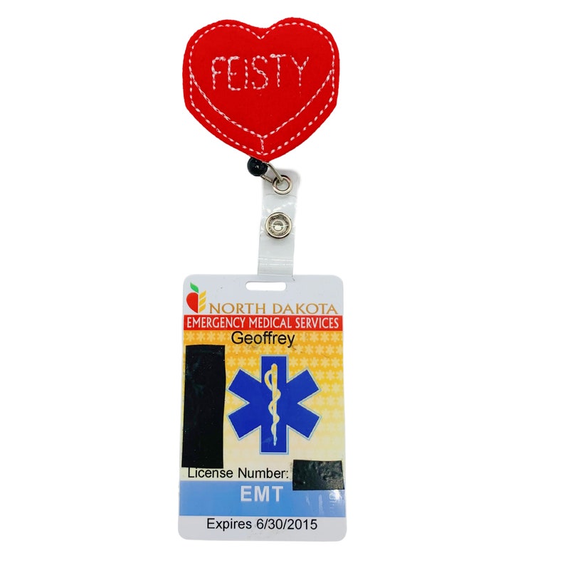 Puede incluir: Un portatarjetas en forma de coraz&oacute;n rojo con la palabra "FEISTY" bordada en blanco. El portatarjetas est&aacute; unido a una tarjeta de identificaci&oacute;n de los Servicios M&eacute;dicos de Emergencia de Dakota del Norte con el nombre "Geoffrey" y una licencia de EMT.