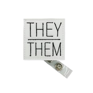 May include: White square badge with black embroidery that reads "THEY/THEM" and a clear plastic badge holder.
