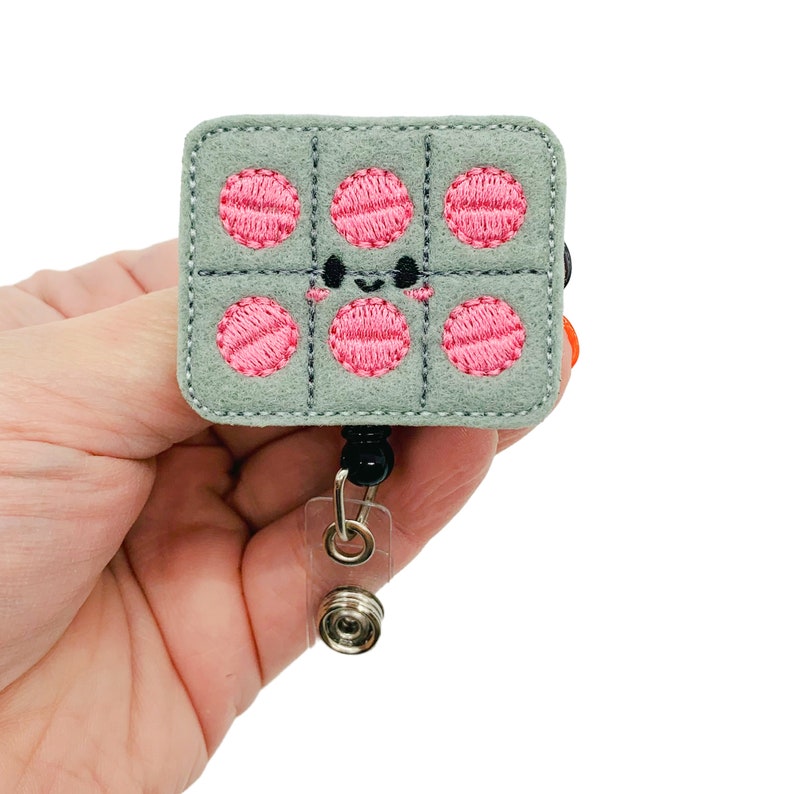 May include: A gray felt badge reel with a cute cartoon waffle design. The waffle has pink circles and a smiling face. The badge reel has a black retractable cord and a clear plastic clip.