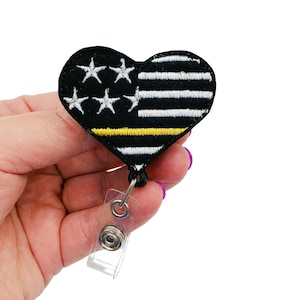 May include: A black heart-shaped badge holder with a thin yellow line and an American flag design. The badge holder has a clear plastic retractable clip.