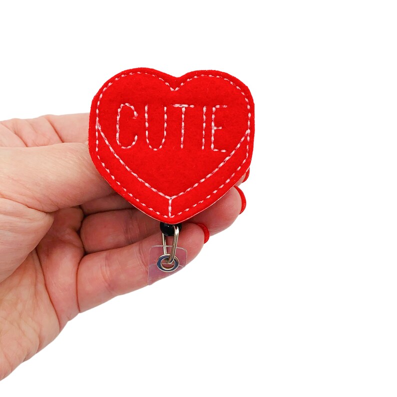 May include: Red felt heart-shaped badge holder with white stitching and the word "CUTIE" embroidered in white thread. The badge holder is attached to a black retractable badge reel.