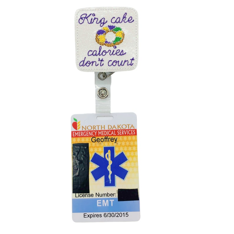 May include: A white badge reel with a King Cake design and the text "calories don't count." The badge reel is attached to a North Dakota Emergency Medical Services ID card with the name "Geoffrey" and an expiration date.