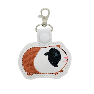 May include: A white and brown guinea pig keychain with a silver clasp. The embroidered design features a guinea pig with a black eye, pink nose, and brown and white fur. The keychain is attached to a white leather backing.