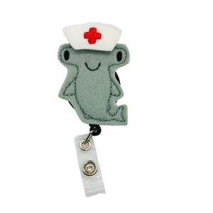 K&ouml;nnte beinhalten: Ein grauer Hammerhai aus Filz, der eine wei&szlig;e Krankenschwesterm&uuml;tze mit einem roten Kreuz tr&auml;gt. Der Hai ist an einem schwarzen, einziehbaren Ausweishalter mit einem durchsichtigen Plastik-Ausweishalter befestigt.