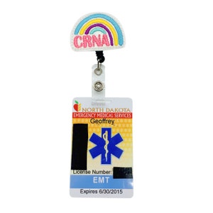 May include: A white badge holder with a rainbow-colored "CRNA" patch and a blue star of life with a snake wrapped around it. The badge holder has the text "NORTH DAKOTA EMERGENCY MEDICAL SERVICES" and "Geoffrey" on it. The badge holder also has the text "License Number: EMT" and "Expires 6/30/2015" on it.