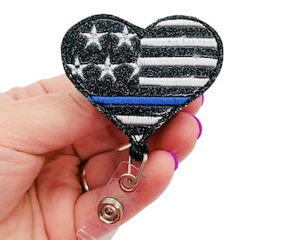 May include: A black and white heart-shaped badge holder with a thin blue line and an American flag design. The badge holder is attached to a clear retractable badge reel.