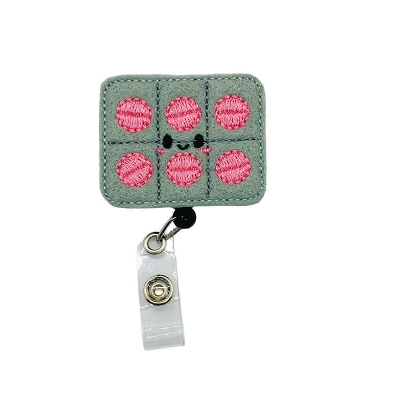 May include: A gray felt badge reel with a smiling face and six pink polka dots. The reel is attached to a clear plastic badge holder.