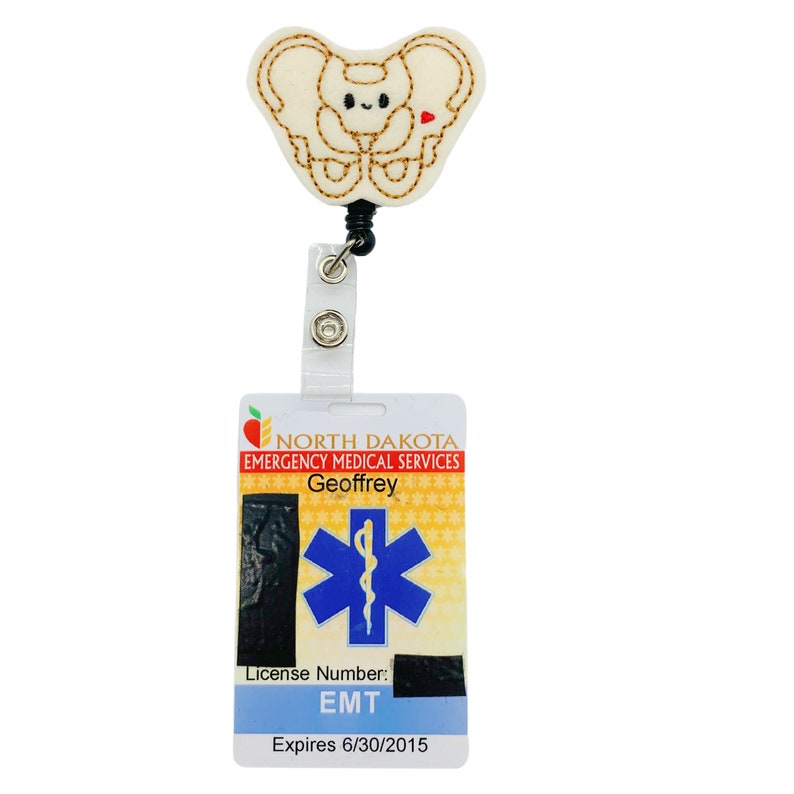 May include: A white felt badge holder with a cartoon image of a pelvis bone with a red heart. The badge holder is attached to a clear plastic retractable badge reel. The badge holder is attached to a white card with the text "NORTH DAKOTA EMERGENCY MEDICAL SERVICES Geoffrey License Number: EMT Expires 6/30/2015". The card also has a blue star of life symbol.