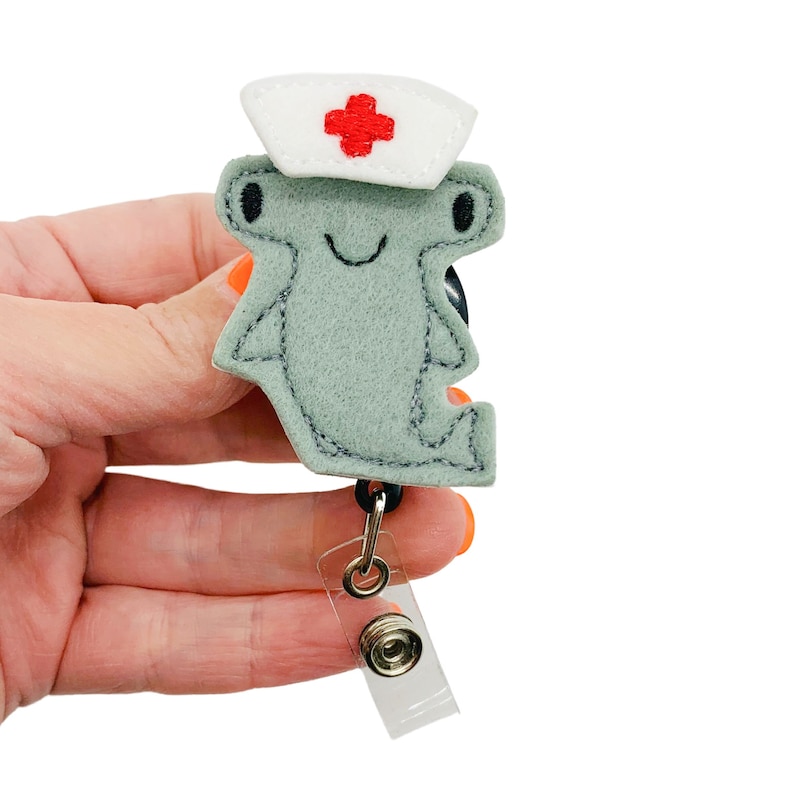K&ouml;nnte beinhalten: Ein grauer Filzhammerhai, der eine wei&szlig;e Krankenschwesterm&uuml;tze mit einem roten Kreuz tr&auml;gt. Der Hai ist an einem durchsichtigen, einziehbaren Ausweishalter aus Kunststoff mit einem silbernen Clip befestigt.