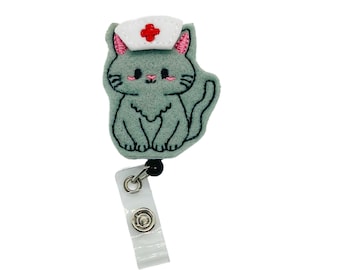 May include: A gray felt cat wearing a white nurse's hat with a red cross. The cat has pink cheeks and black eyes. The cat is attached to a clear plastic retractable badge holder.