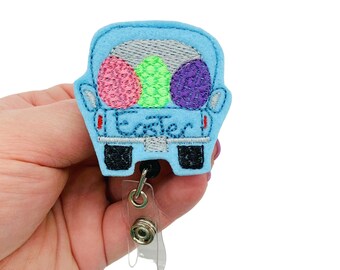 May include: A light blue felt badge reel featuring a truck bed filled with embroidered Easter eggs in pink, green, and purple. The word "Easter" is embroidered on the truck. The badge reel has a retractable cord and a clear plastic clip.