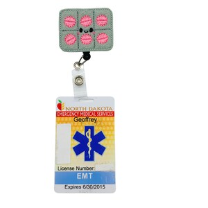 May include: A light blue and yellow ID badge with the text "NORTH DAKOTA EMERGENCY MEDICAL SERVICES" and "Geoffrey". The badge also features a blue star of life with a snake wrapped around it. The badge has the text "License Number: EMT Expires 6/30/2015". A gray and pink pill-shaped badge holder with a smiling face is attached to the ID badge.