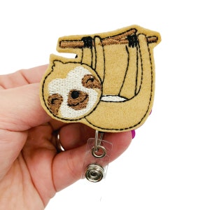 May include: A brown and white embroidered sloth hanging from a branch with a clear plastic retractable badge holder attached.
