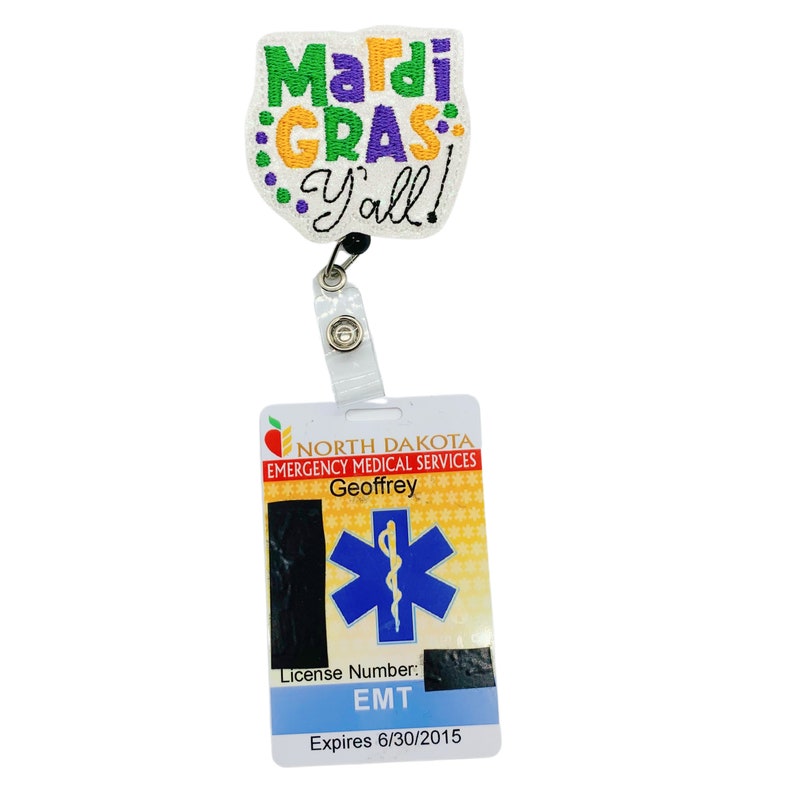 May include: A white badge holder with a Mardi Gras Y'all! embroidered patch attached. The badge holder has a clear plastic strap and a metal clip. The badge itself is white with a red apple logo, the text "NORTH DAKOTA EMERGENCY MEDICAL SERVICES", and the name "Geoffrey". It also features a blue star of life with a snake wrapped around it. The badge has the text "License Number: EMT" and "Expires 6/30/2015".
