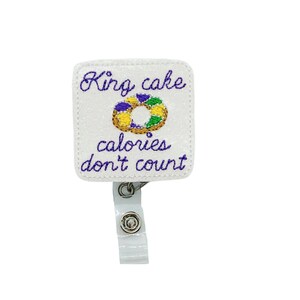May include: A white, square badge reel with the embroidered words "King cake calories don't count" in purple script. The design includes a colorful embroidered king cake. The reel has a clear plastic strap and a metal clip.