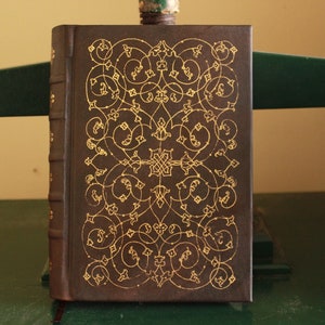 K&ouml;nnte beinhalten: Ein braunes, ledergebundenes Buch mit einem aufwendigen goldenen Design auf dem Einband.
