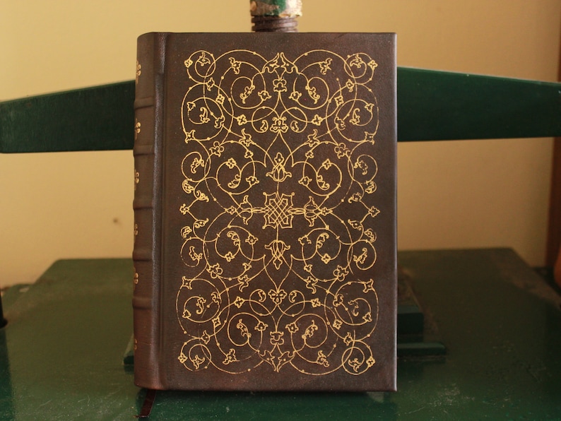 K&ouml;nnte beinhalten: Ein braunes, ledergebundenes Buch mit einem aufwendigen goldenen Design auf dem Einband.