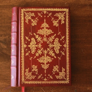 May include: A red leather-bound book with a gold-colored intricate design on the cover.