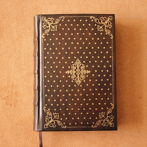Op de afbeelding: Een donkerbruin leren notitieboek met gouden ster- en bloemaccenten. De kaft heeft een centraal sierlijk ontwerp en hoekversieringen. Een lint-bladwijzer steekt uit de rug.