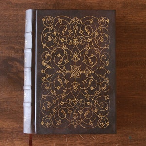 K&ouml;nnte beinhalten: Ein dunkelbraunes, ledergebundenes Tagebuch mit einem aufwendigen goldenen Blumendesign auf dem Einband.