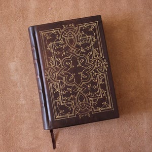 Puede incluir: Un diario de cuero marrón oscuro con un diseño de nudos celtas dorados en relieve en la portada. El lomo tiene bandas en relieve y una cinta de marcador marrón visible. El diario está sobre una superficie marrón.