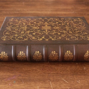 K&ouml;nnte beinhalten: Ein braunes, ledergebundenes Buch mit goldgepr&auml;gten floralen Mustern auf dem Einband. Das Buch hat f&uuml;nf erhabene B&auml;nder auf dem R&uuml;cken.