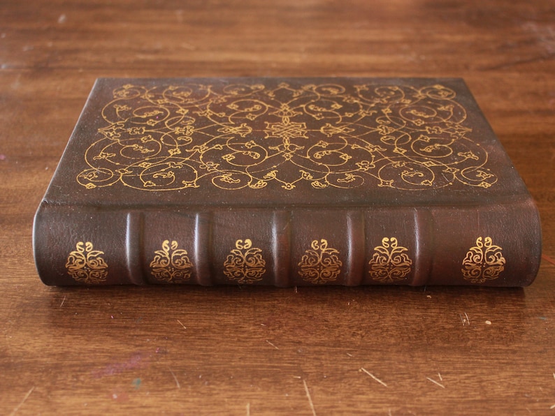 K&ouml;nnte beinhalten: Ein braunes, ledergebundenes Buch mit goldgepr&auml;gten floralen Mustern auf dem Einband. Das Buch hat f&uuml;nf erhabene B&auml;nder auf dem R&uuml;cken.